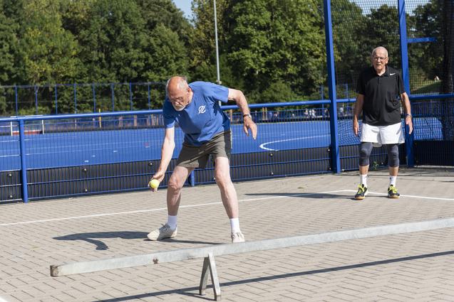 Zwei Männer auf einem Sportplatz, einer schlagt einen Ball, der andere steht wartend im Hintergrund.