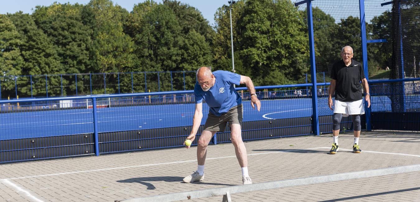Zwei Männer auf einem Sportplatz, einer schlagt einen Ball, der andere steht wartend im Hintergrund.