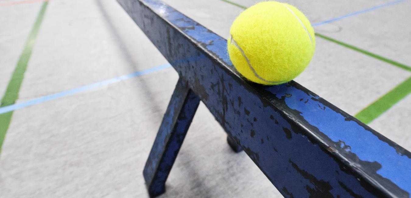 Gelber Tennisball liegt auf einer blauen Metallstange in einer Sporthalle mit farbigen Linien auf dem Boden.