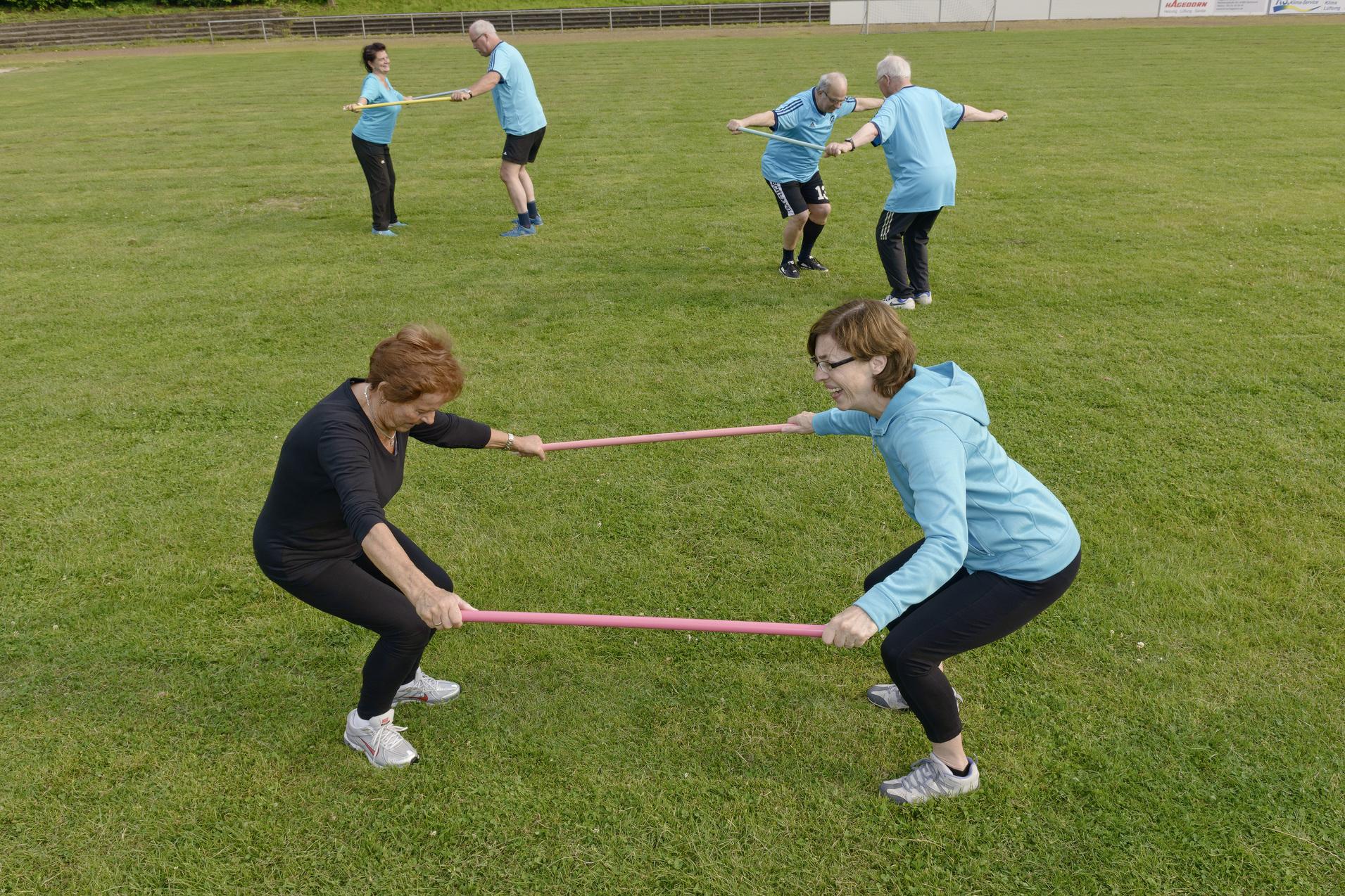 Zwei Frauen ziehen in einem Fitness&uuml;bung auf einem Rasen mit einem Widerstandsband, w&auml;hrend andere im Hintergrund aktiv sind.