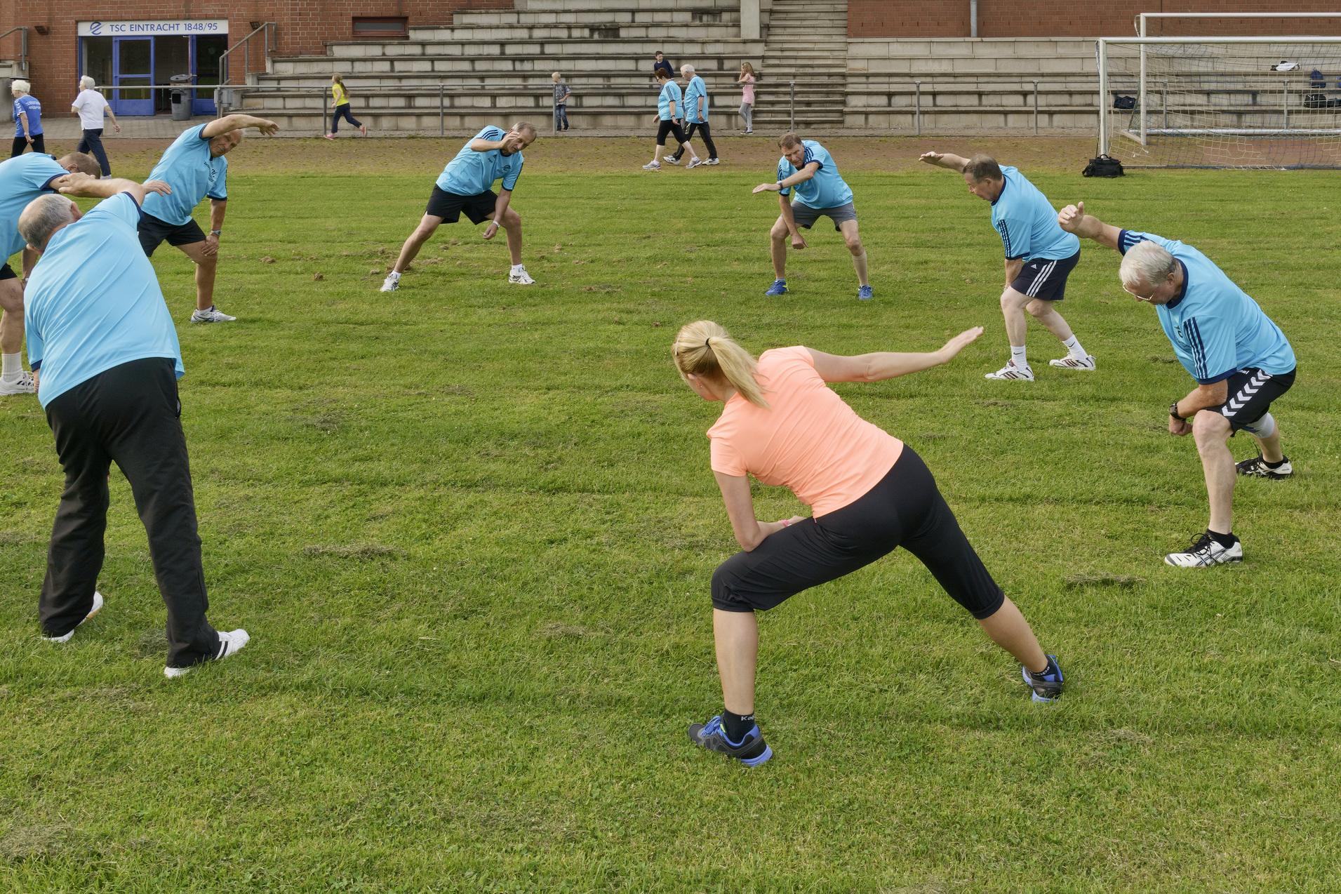 Gruppe von Menschen in Sportkleidung beim Dehnen auf einem gr&uuml;nen Rasenfeld.