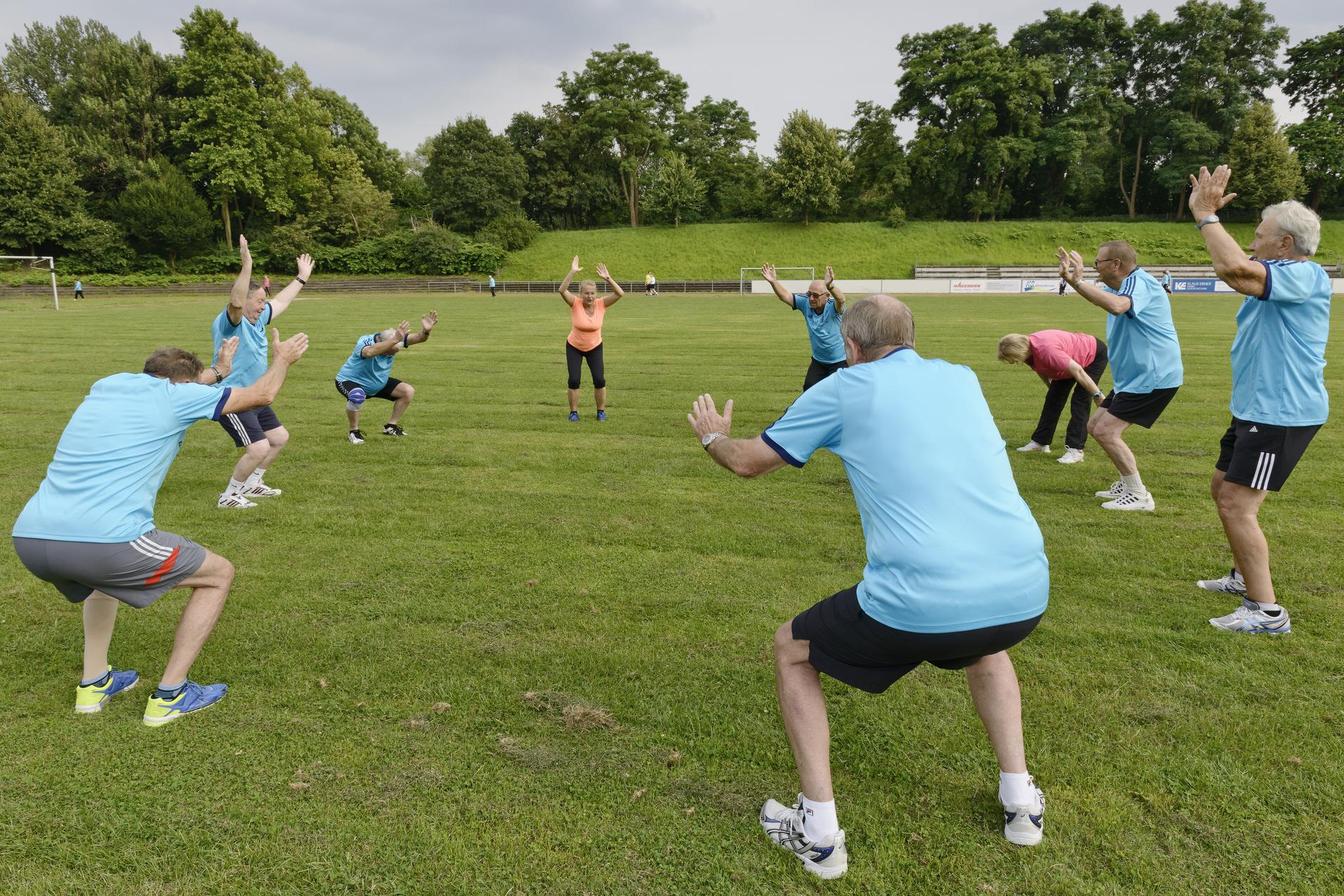 Gruppe &auml;lterer Menschen in blauen T-Shirts bei einem Outdoor-Fitnesskurs auf einem gr&uuml;nen Sportplatz.