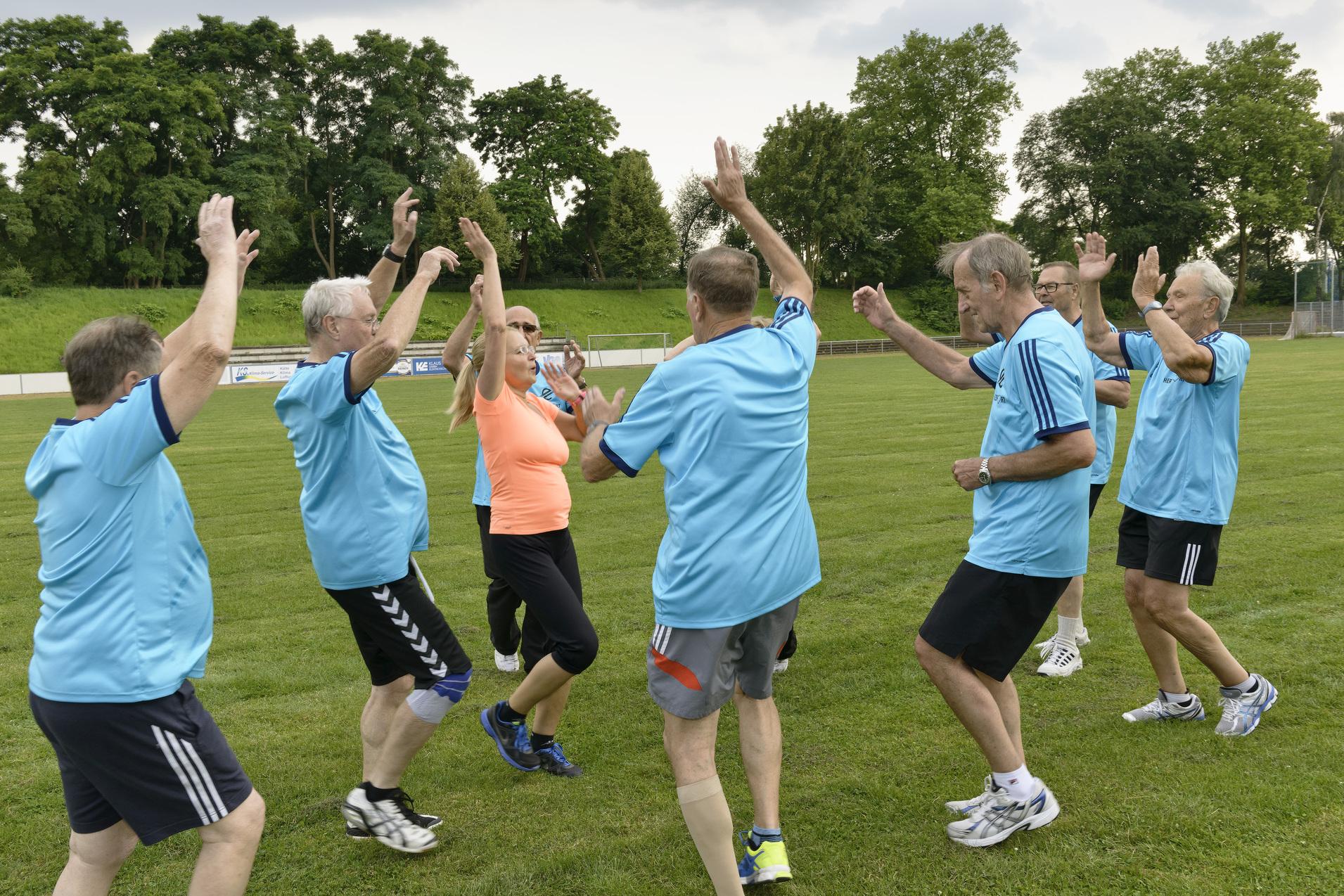 Gruppe von &auml;lteren M&auml;nnern in blauen T-Shirts tanzen auf einem Fu&szlig;ballfeld, umgeben von B&auml;umen und Himmel.