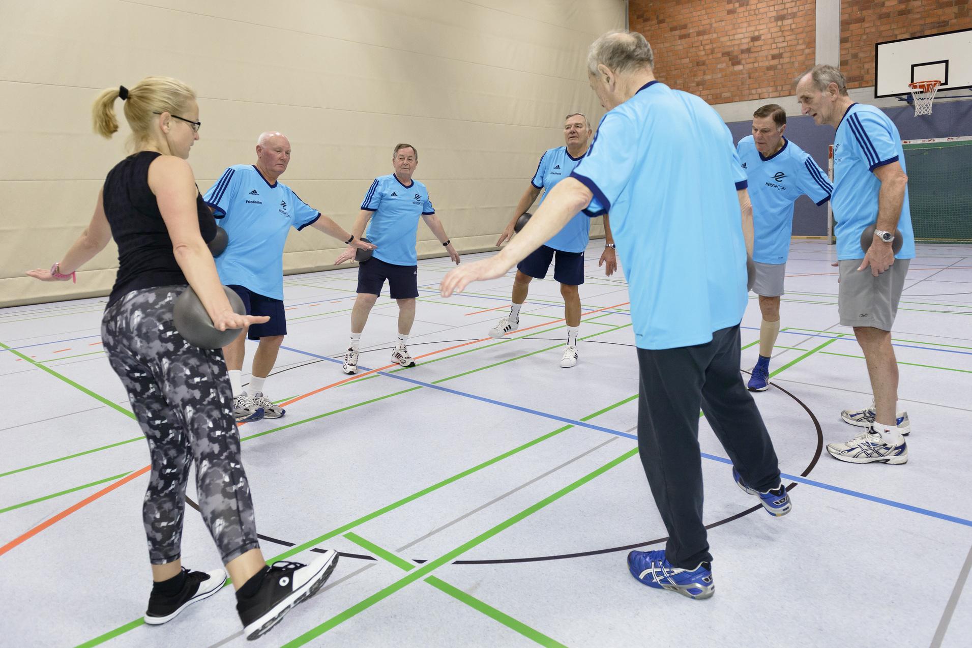 Gruppe von sechs &auml;lteren M&auml;nnern in blauen T-Shirts und einer Frau in Leggings beim Training in einer Sporthalle.
