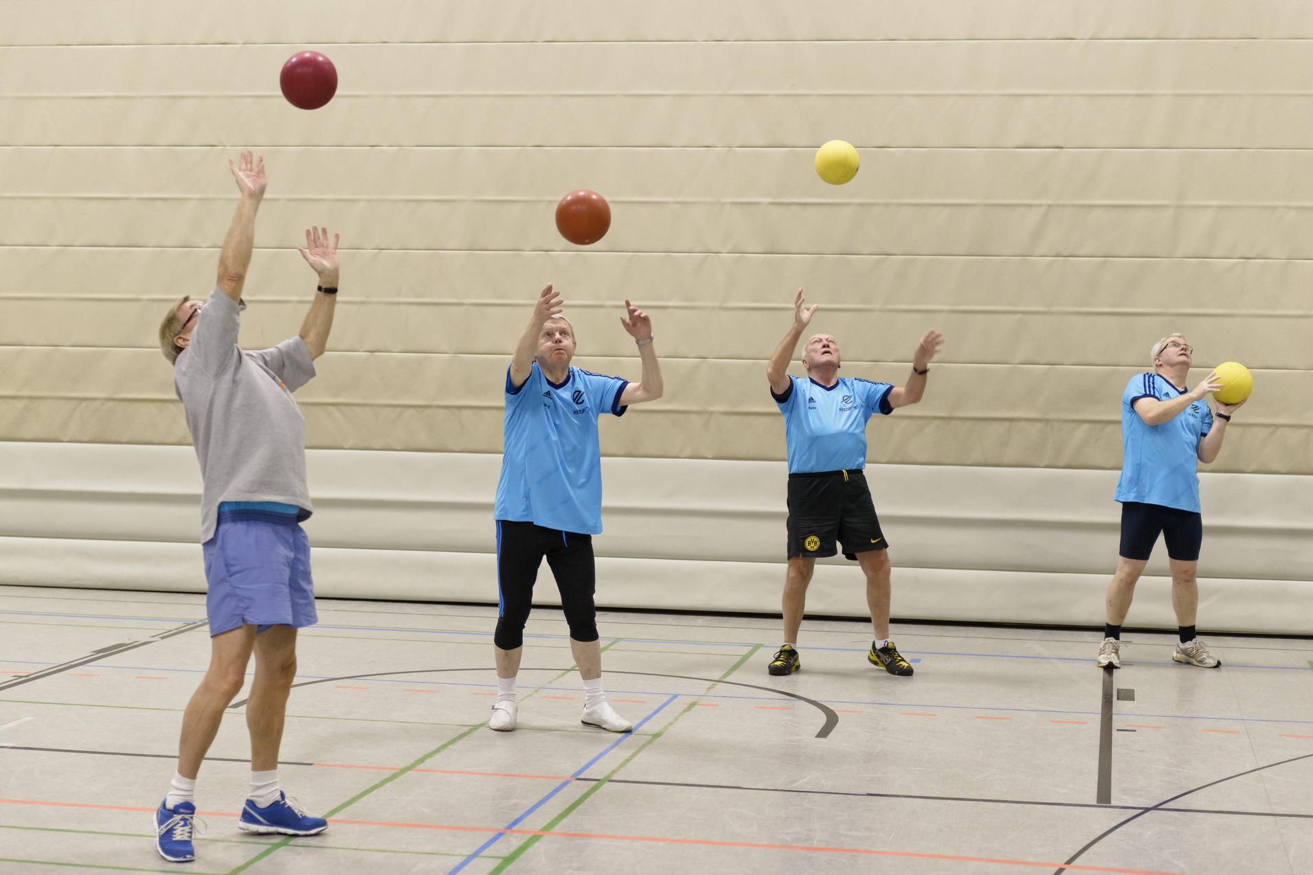 Vier M&auml;nner in blauen T-Shirts werfen B&auml;lle in einer Sporthalle in die Luft.