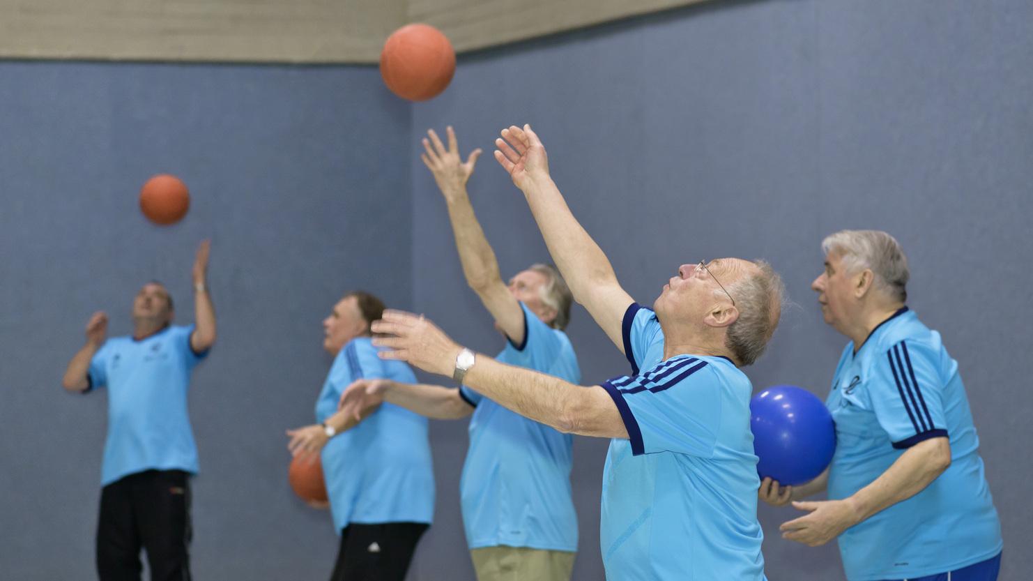 Gruppe &auml;lterer M&auml;nner in blauen T-Shirts spielt mit B&auml;llen in einer Sporthalle.
