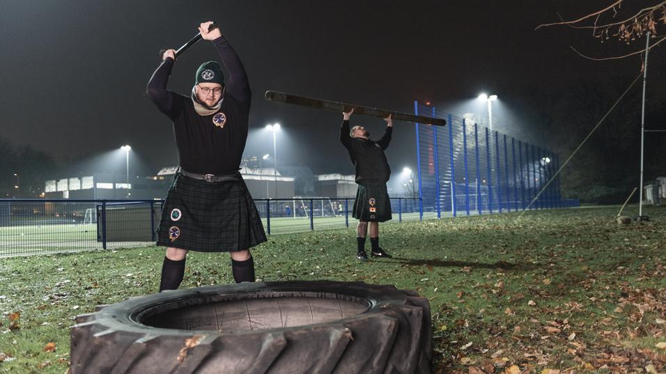 Zwei M&auml;nner in Kilts trainieren im Dunkeln mit einer Reifenheber-&Uuml;bung und einem Holzstamm auf einem Sportplatz.