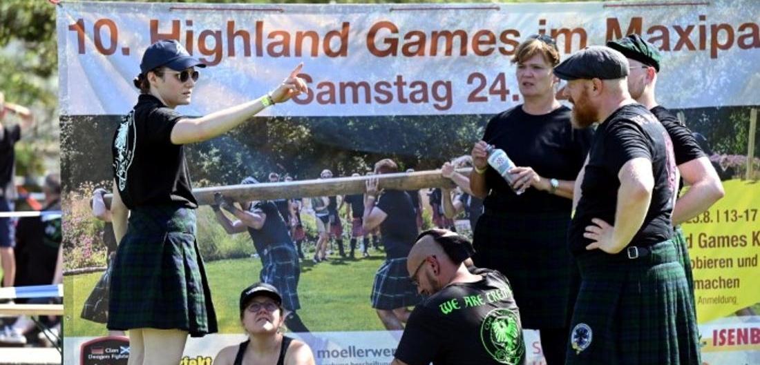 Gruppe von Menschen in Schottenröcken bei den Highland Games, Diskussion vor einem Veranstaltungsplakat im Freien.