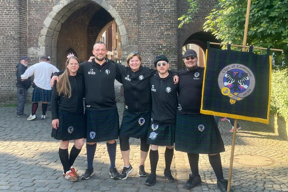 Gruppe von fünf Personen in schwarzen T-Shirts und Kilts steht vor einer historischen Torbogen-Architektur.