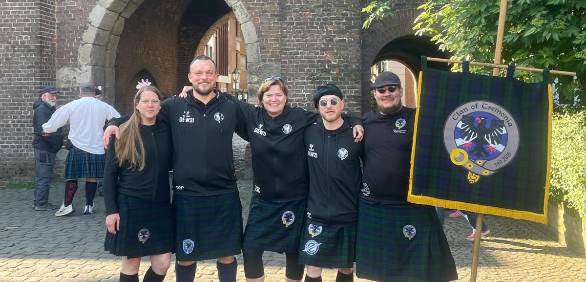 Gruppe von fünf Personen in schwarzen T-Shirts und Kilts steht vor einer historischen Torbogen-Architektur.