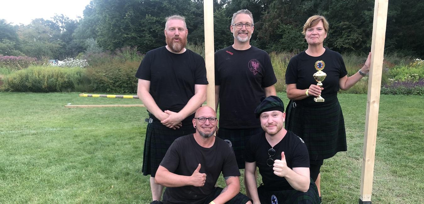Fünf Personen in schottischen Kilts posieren auf einer Wiese, eine hält einen Pokal in der Hand.