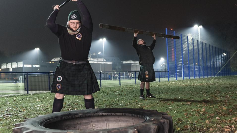 Zwei M&auml;nner in Kilts trainieren im Dunkeln mit einer Reifenheber-&Uuml;bung und einem Holzstamm auf einem Sportplatz.