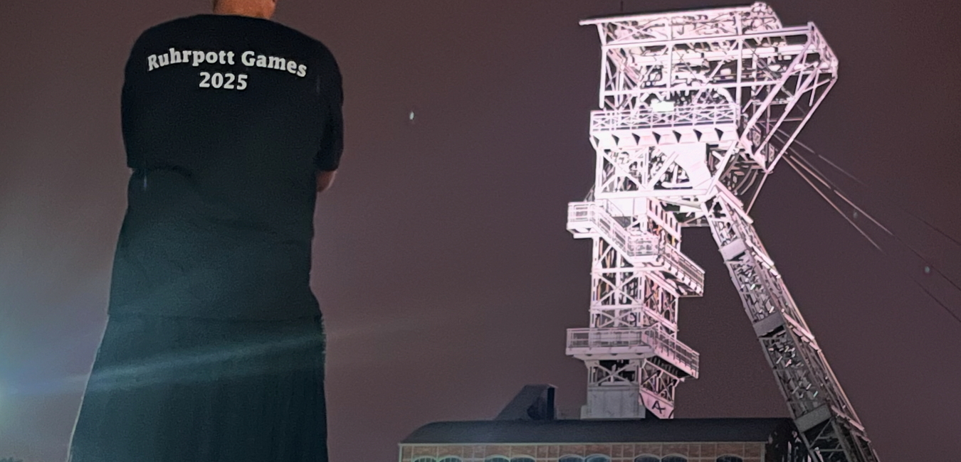 Person mit T-Shirt &bdquo;Ruhrpott Games 2025&ldquo; steht vor einer beleuchteten F&ouml;rderturmkonstruktion bei Nacht.