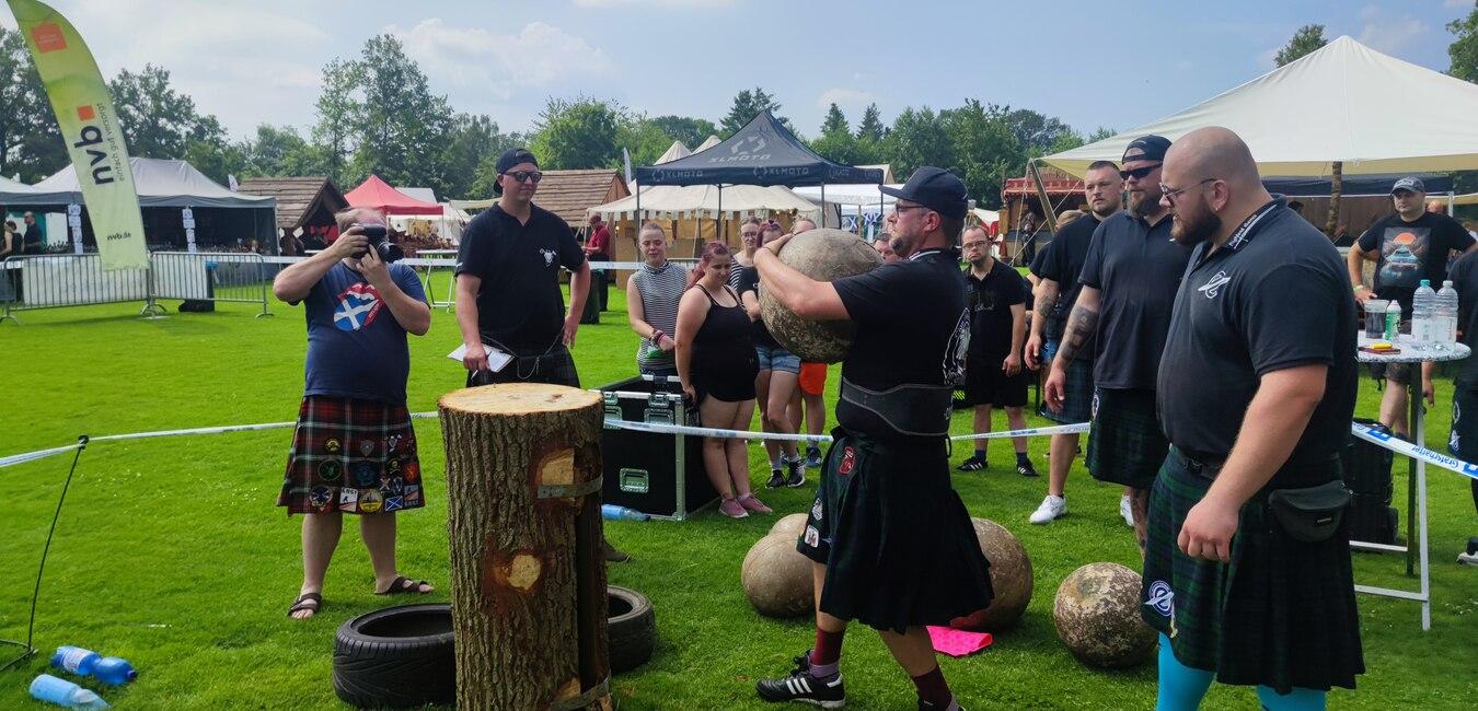 Menschen in schottischer Tracht treten bei einem Wettkampf auf einer grünen Wiese gegeneinander an.