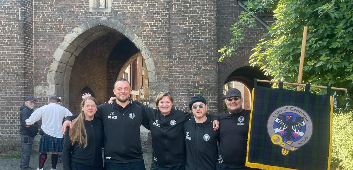 Gruppe von fünf Personen in schwarzen T-Shirts und Kilts steht vor einer historischen Torbogen-Architektur.