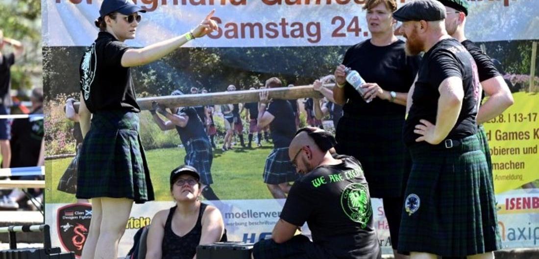 Gruppe von Menschen in Schottenröcken bei den Highland Games, Diskussion vor einem Veranstaltungsplakat im Freien.