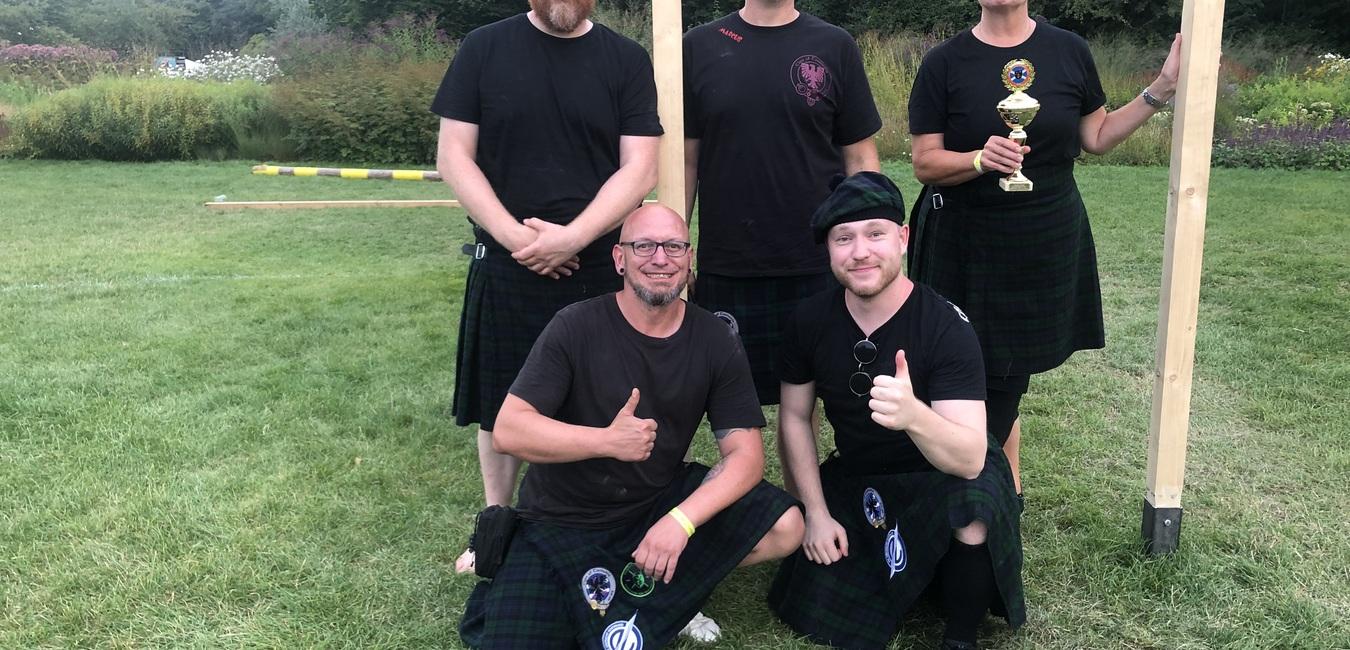 Fünf Personen in schottischen Kilts posieren auf einer Wiese, eine hält einen Pokal in der Hand.
