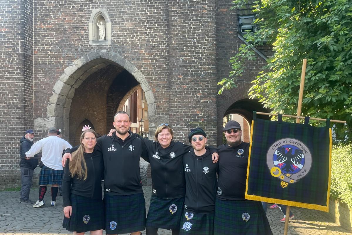 Gruppe von f&uuml;nf Personen in schwarzen T-Shirts und Kilts steht vor einer historischen Torbogen-Architektur.