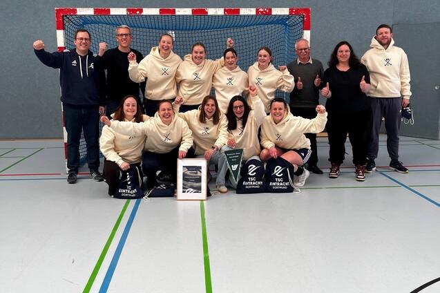 Gruppe von Handballspielern in wei&szlig;en Sweatshirts posiert vor einem Tor, l&auml;chelnd mit Pokalen und Taschen.