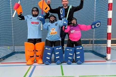 Gruppe von vier Personen in Sportausr&uuml;stung, die in einem Hockeytor posieren, ein Trainer hebt jubelnd die Arme.