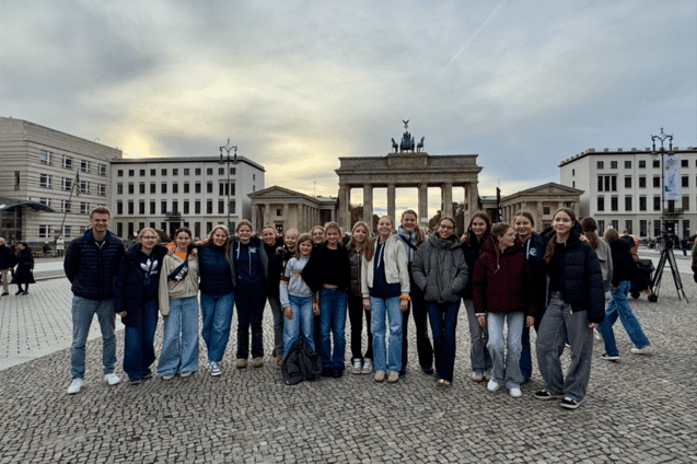 Gruppe von jungen Erwachsenen posiert l&auml;chelnd vor dem Brandenburger Tor, umgeben von historischen Geb&auml;uden.