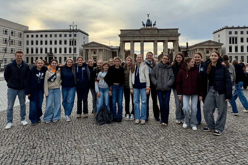 Gruppe von jungen Erwachsenen posiert l&auml;chelnd vor dem Brandenburger Tor, umgeben von historischen Geb&auml;uden.