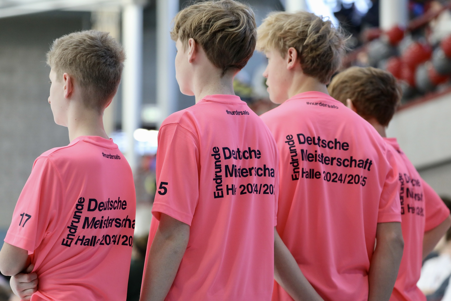 Vier Jungs in pinken Trikots stehen nebeneinander, mit dem R&uuml;cken zur Kamera, im Hintergrund eine Sporthalle.