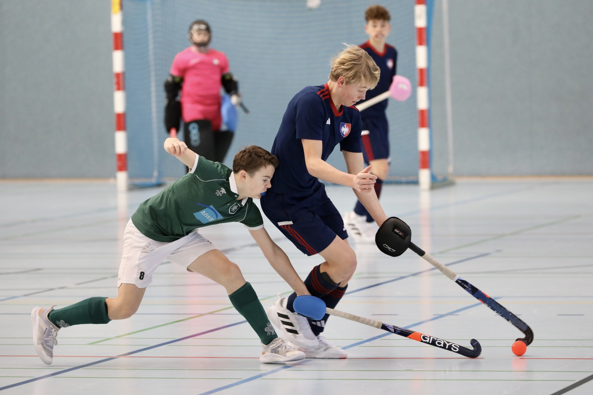 Zwei Jugendliche spielen Hockey in einer Sporthalle, einer versucht, den Ball von einem anderen Spieler zu stehlen.