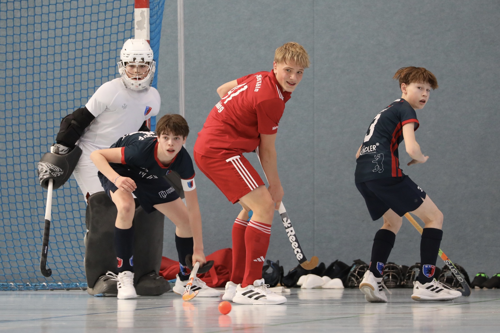 Drei junge Hockeyspieler auf einem Sportboden, ein Torwart in der Ecke, zwei Spieler in Aktion mit einem Ball.