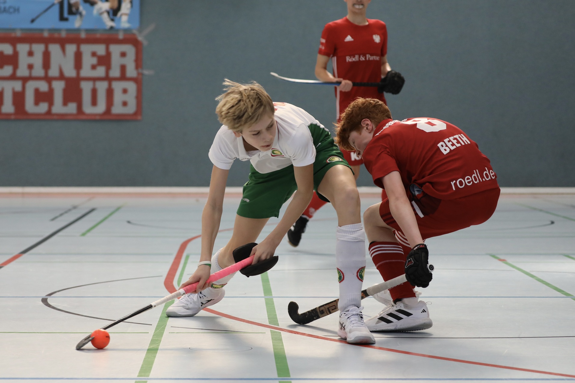 Zwei junge Spieler beim Hockeyspiel in einer Sporthalle, einer mit wei&szlig;em und gr&uuml;nem Trikot, der andere in rotem Trikot.