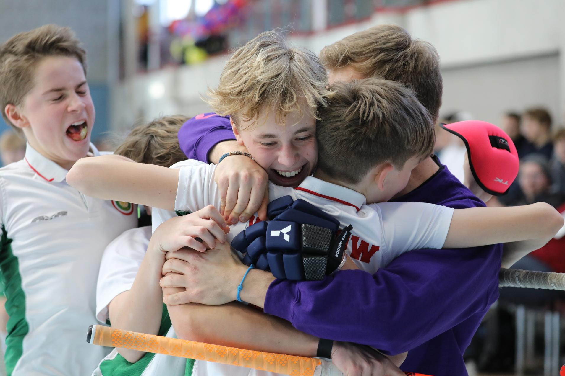 Gruppe jubelnder Kinder umarmt sich nach einem Sieg, mit Hockeyschl&auml;gern und Sportkleidung, in einer Sporthalle.