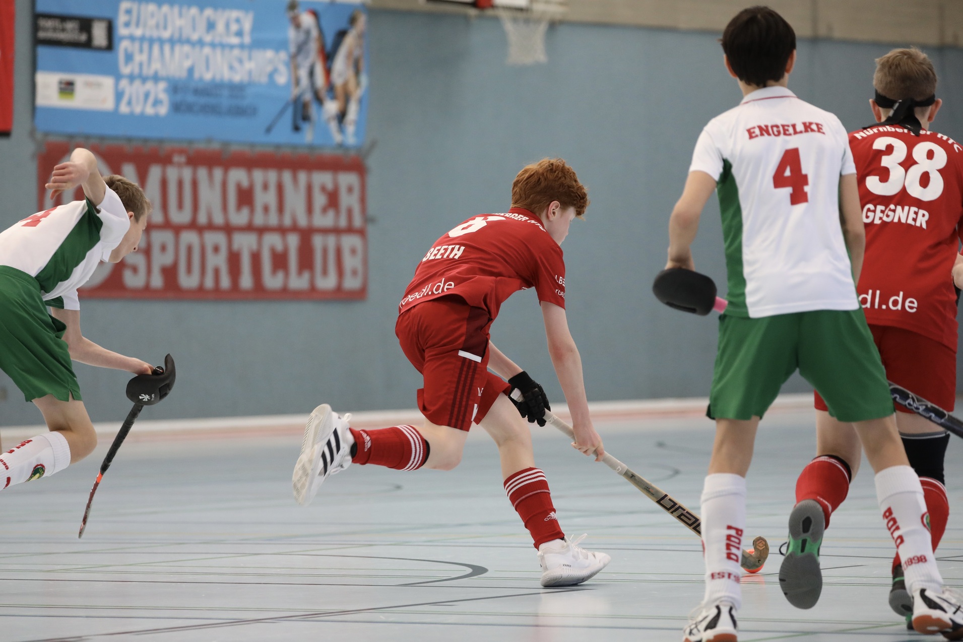 Zwei Gruppen von Jugendlichen spielen Hallenhockey, einer in rotem, der andere in gr&uuml;nem Trikot, auf einem Sportboden.