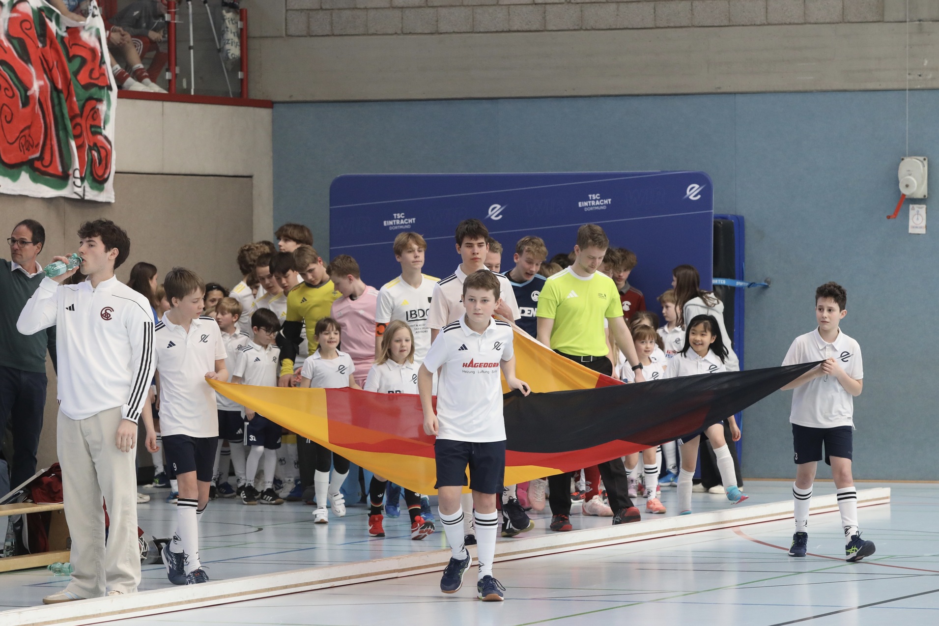 Kinder in Sportkleidung tragen eine Deutschlandflagge und betreten eine Sporthalle. Zuschauer im Hintergrund.