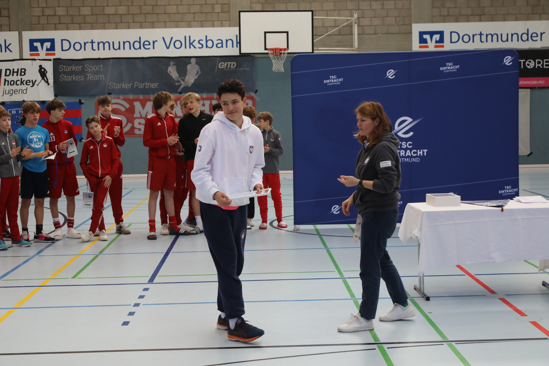 Junger Sportler erh&auml;lt eine Auszeichnung von einer Frau, w&auml;hrend eine Gruppe Kinder im Hintergrund zuschaut.