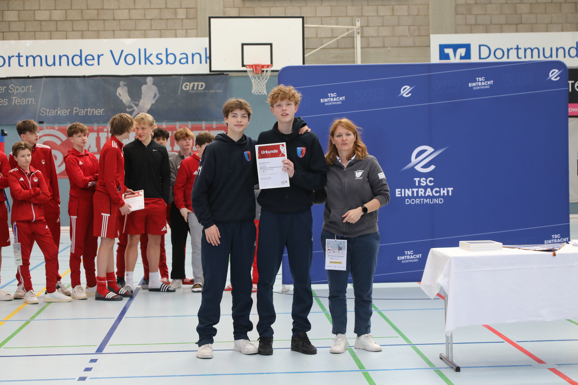 Zwei Jugendsportler stehen mit Urkunde vor einer Gruppe von Spielern in roten Trainingsanz&uuml;gen, w&auml;hrend eine Trainerin neben ihnen steht.