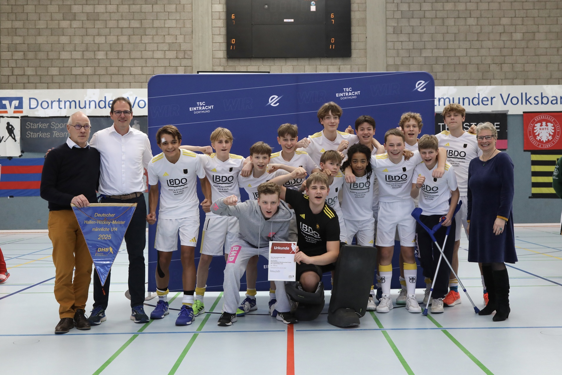 Gruppe von Jugendspielern posiert nach einem Turnier, umgeben von Trainern und einer Ehrentafel in einer Sporthalle.