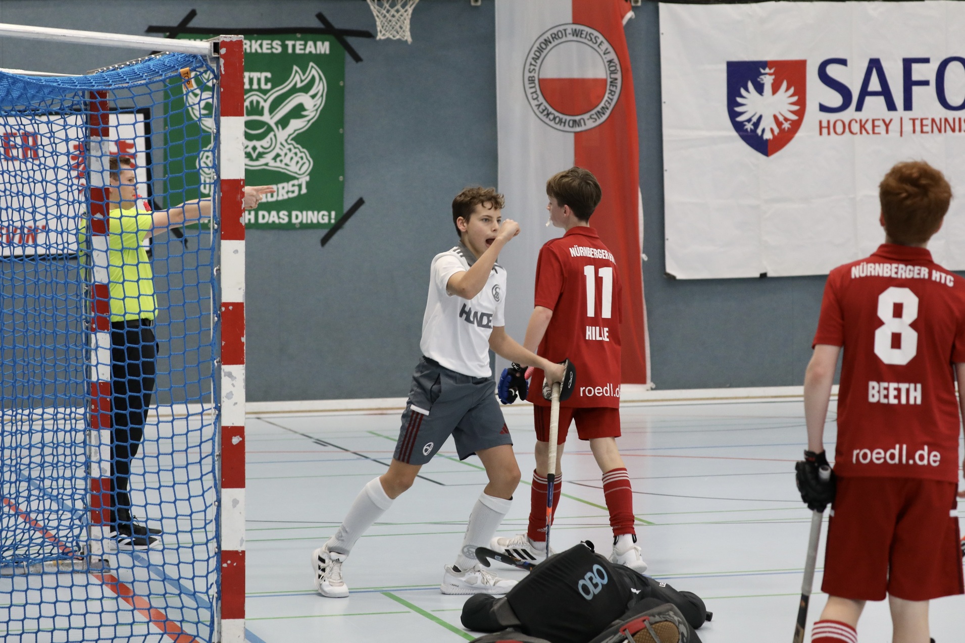 Zwei Kinder in Sportkleidung spielen Hockey in einer Halle, einer jubelt gegen&uuml;ber einem Mitspieler in rotem Trikot.