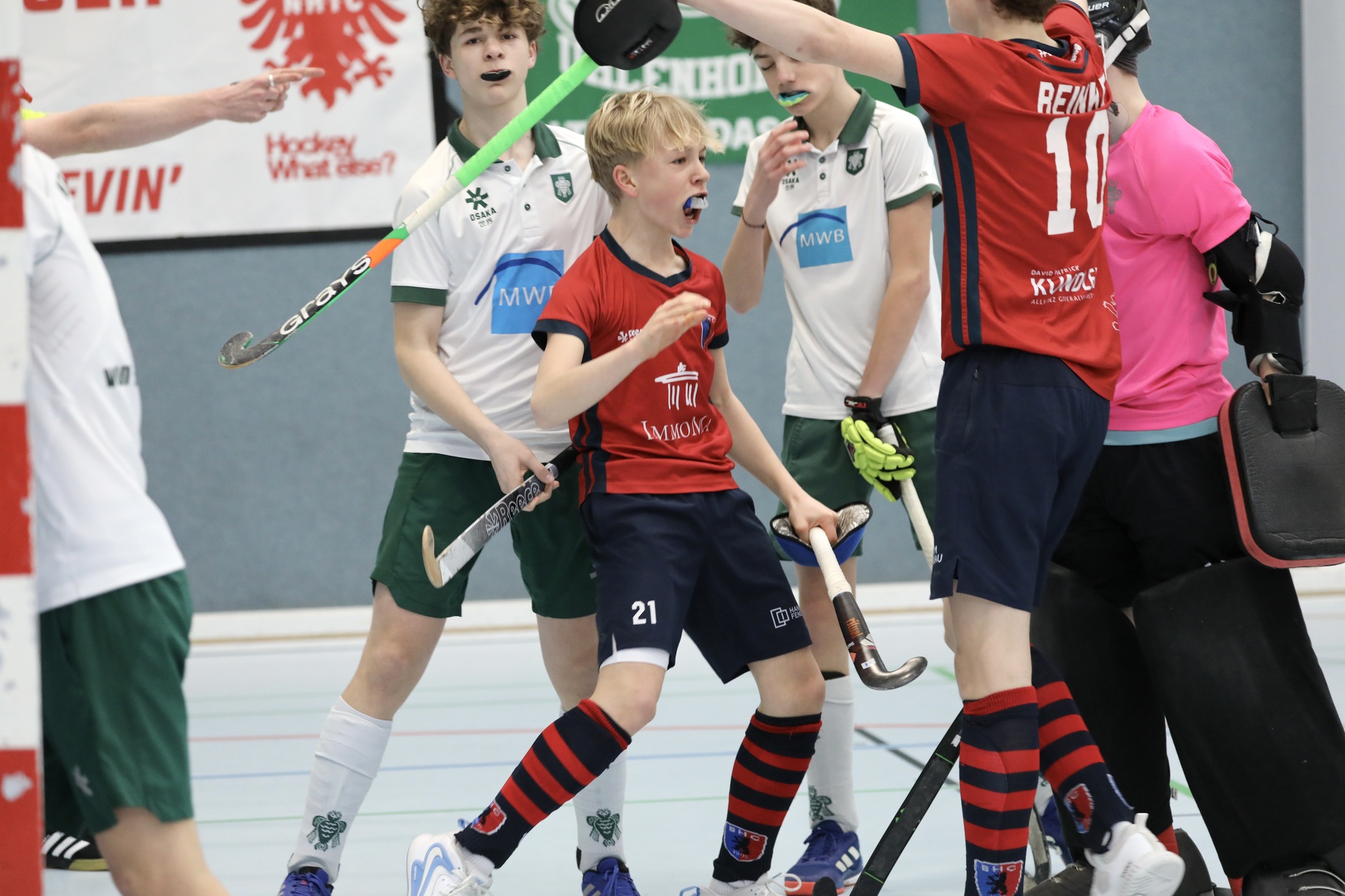 Jubelnde Spieler in rot und schwarz feiern ein Tor in einem Hallenhockeyspiel. Hintergrund mit anderen Spielern in Wei&szlig; und Gr&uuml;n.