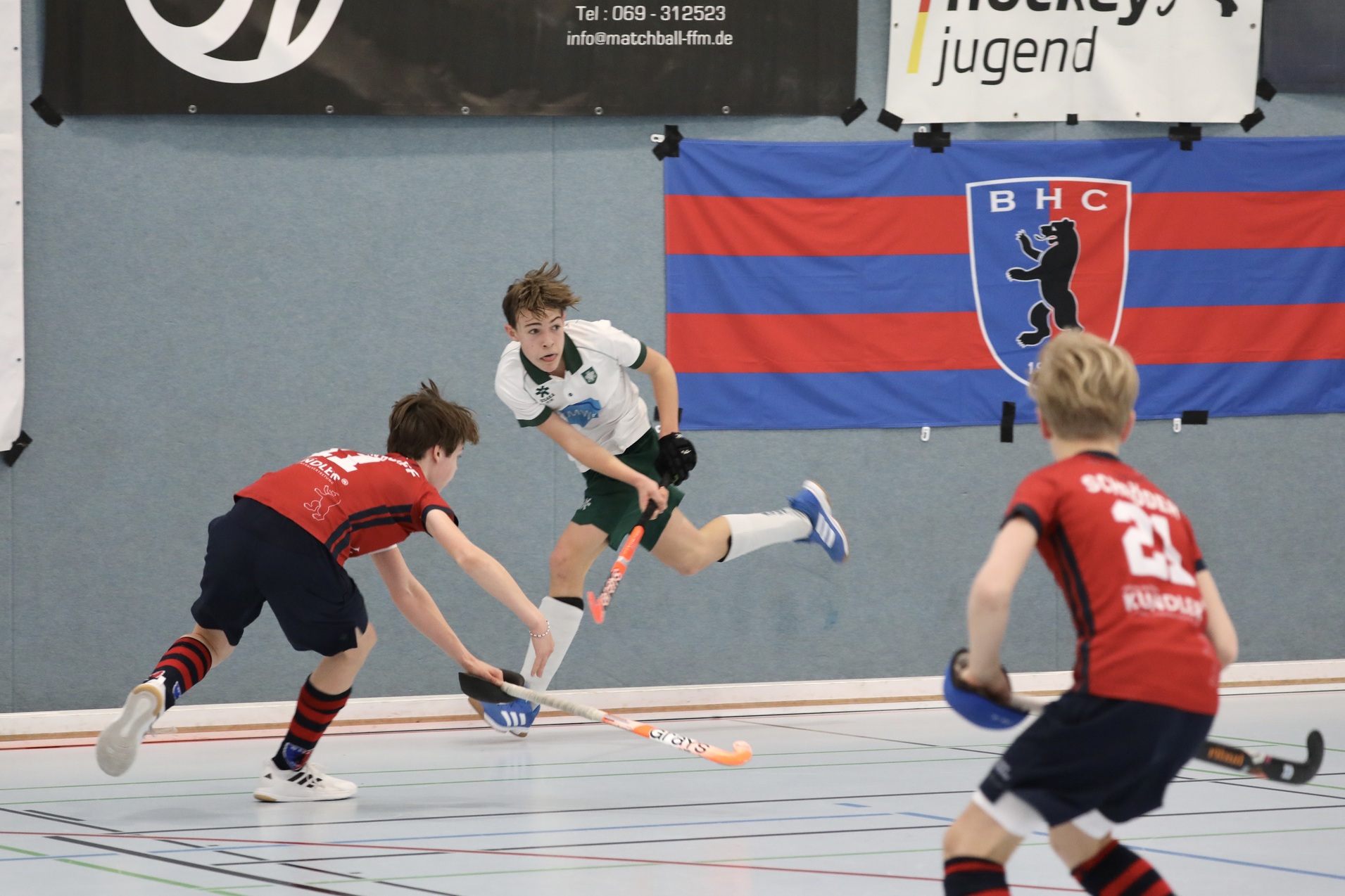 Zwei Kinder spielen in einer Sporthalle Hockey; ein Spieler in Wei&szlig; hebt seinen Schl&auml;ger, w&auml;hrend ein Gegenspieler angreift.