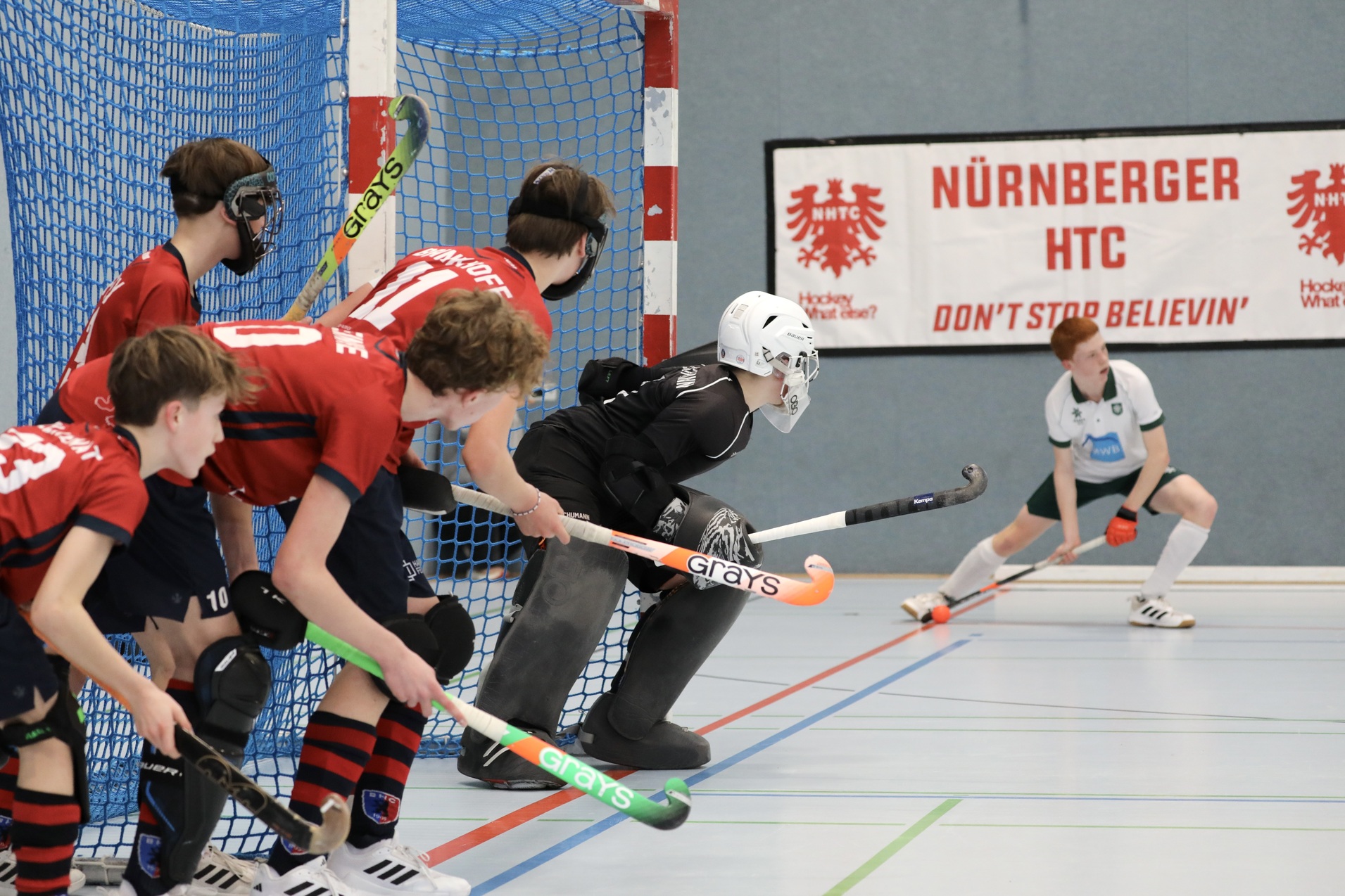 Jugendliche Hockeyspieler in roten Trikots stehen vor dem Torwart in schwarzer Ausr&uuml;stung und einer bunten Bande.