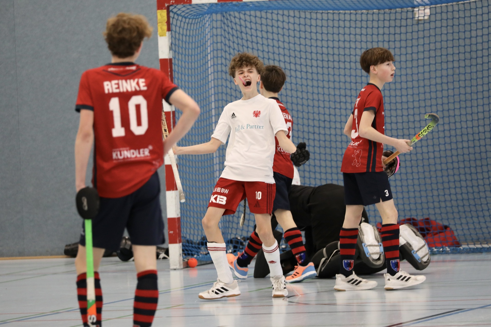 Junge Spieler in Sportkleidung feiern einen Treffer in einer Hallenhockeypartie, im Hintergrund weitere Spieler.