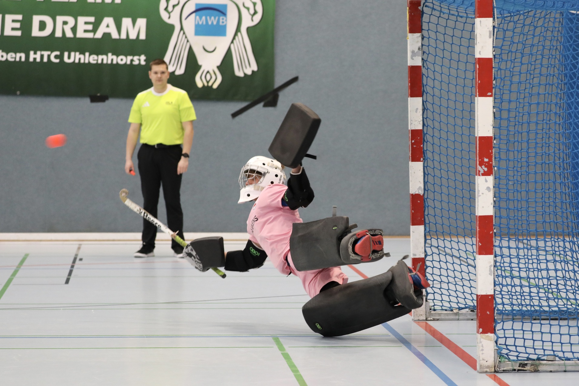 Torwart in pinkem Trikot springt zur Seite, um einen Ball w&auml;hrend eines Hallenhockeyspiels abzuwehren. Schiedsrichter im Hintergrund.