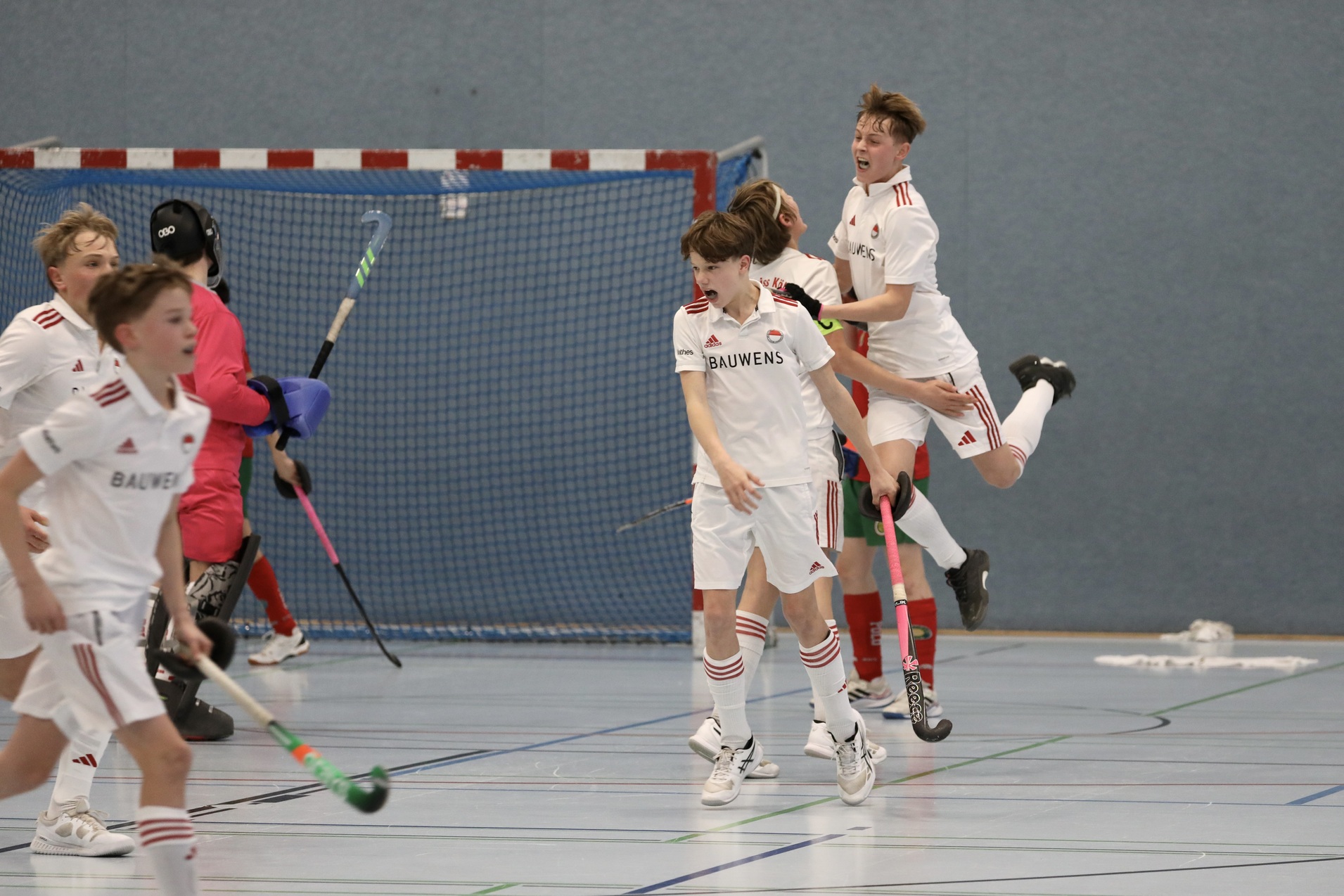 Junge Hockeyspieler in wei&szlig;en Trikots feiern ein Tor in einer Sporthalle mit blauem Hintergrund.