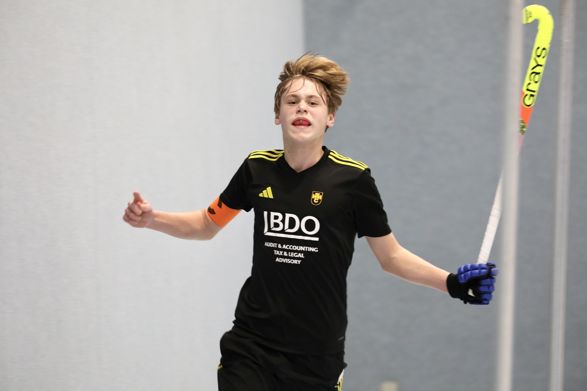 Junger Hockeyspieler in schwarzem Trikot jubelt mit einem Schl&auml;ger in der Hand auf einem Hallenboden.