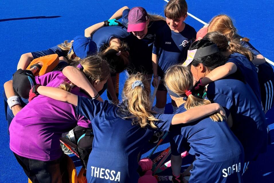 M&auml;dchenteam umarmt sich im Kreis auf einem blauen Hockeyfeld, im Hintergrund sind andere Spieler und Zelte sichtbar.