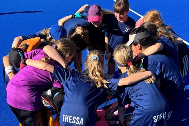 M&auml;dchenteam umarmt sich im Kreis auf einem blauen Hockeyfeld, im Hintergrund sind andere Spieler und Zelte sichtbar.