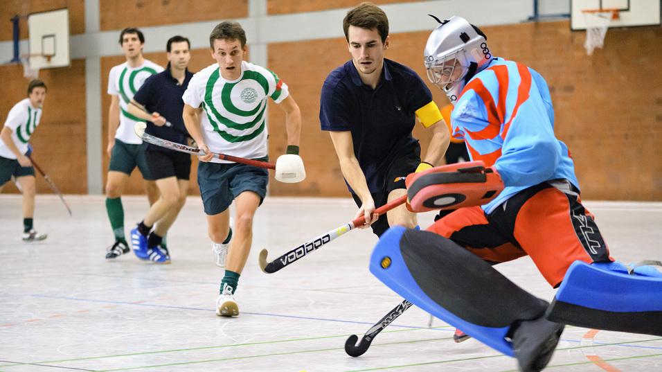 Mehrere Spieler in einem Hallenhockeyspiel, ein Torwart weicht einem Schuss in die Ecke aus. Gelbe Hockeykugel sichtbar.