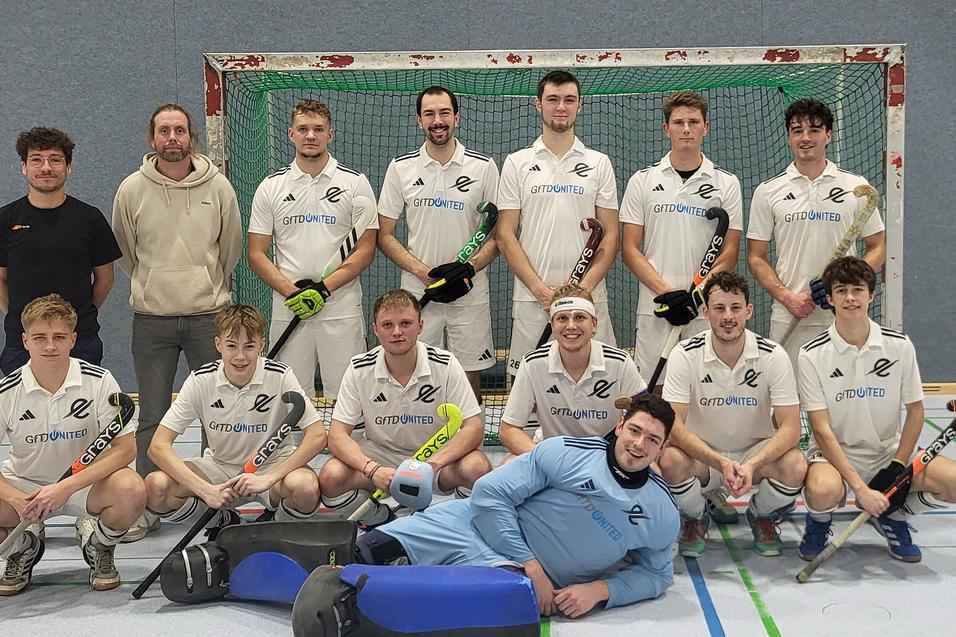 Mannschaftsfoto einer Hockeymannschaft in wei&szlig;en Trikots, aufgenommen vor einem Tor in einer Sporthalle.