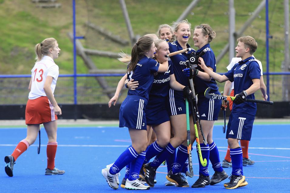 Gruppe junger Hockeyspielerinnen jubelt auf dem Spielfeld, w&auml;hrend eine Spielerin in Wei&szlig; im Hintergrund steht.