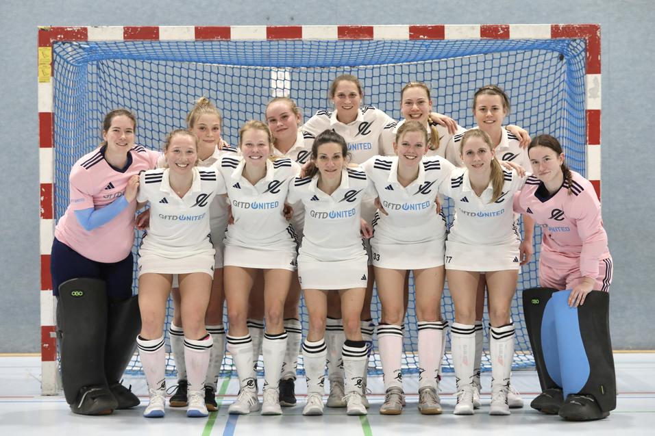 Frauen-Handballmannschaft posiert l&auml;chelnd vor einem Tor in der Halle, einheitlich in wei&szlig;en Trikots gekleidet.